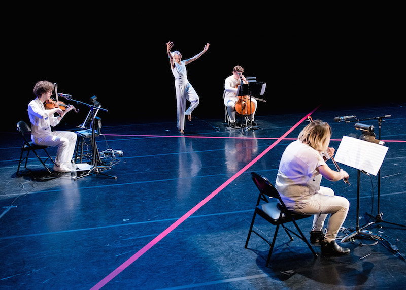 McIntyre dressed in white reaching her arms overhead while the string trio sits in chairs around her.Thick pieces of pink tape make an x on the stage